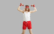 Funny thin nerd with dumbbells exercising weak muscles isolated on gray background. Hilarious smiling young man in eyeglasses, retro headband, white tank top and red shorts enjoying his sports workout