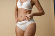 © Kostiantyn - Body of mature woman in underwear touching her skin, holding hand on waist, standing isolated over beige background