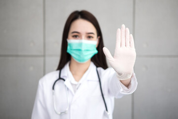 Asian woman doctor who wears medical coat and face mask shows hand as “stop” sign in health protection concept at hospital. Stop epidemic of COVID-19.