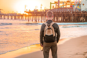 View from the back of a young blonde man with a backpack on Santa Monica beach against the background of an orange sunset in Los Angeles, California