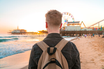 View from the back of a young blonde man with a backpack on Santa Monica beach against the background of an orange sunset in Los Angeles, California