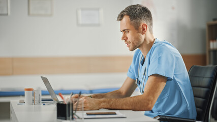 Wall Mural - Experienced Male Nurse Wearing Blue Uniform Working on Laptop Computer at Doctor's Office. Medical Health Care Professional Working On Battling Stereotypes to Gender Diversity in Nursing Career.