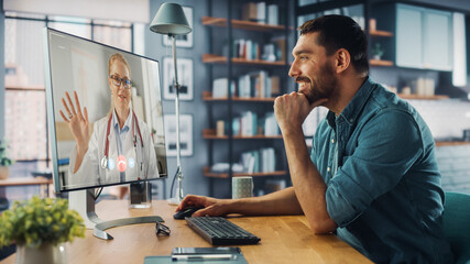 Wall Mural - Handsome Caucasian Male is Making a Video Call to His Medical Consultant on Desktop Computer at Home Living Room while Sitting at Table. Man Working From Home and Talking to a Doctor Over the Internet