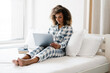 © Drobot Dean - The smiling black woman sitting on the bed in front of laptop near window