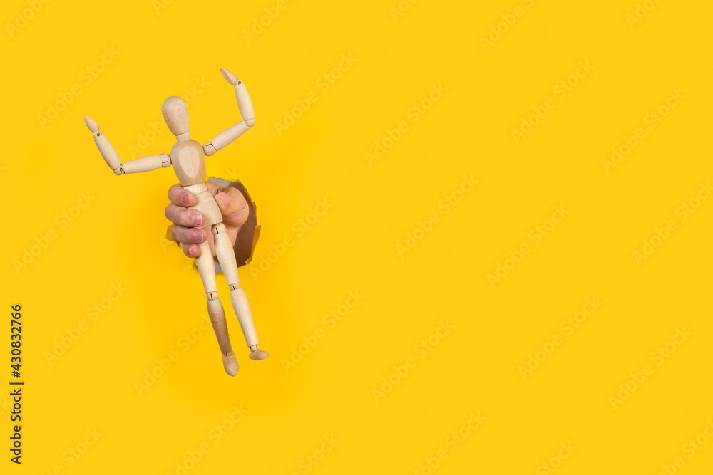 Mano de hombre sujetando la figura de madera modelo maniquí a travéz de un agujero de un papel amarillo liso y aislado. Vista de frente. Copy space - obrazy, fototapety, plakaty 