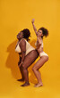 © Svitlana - Full length shot of two funny slim and plus size african american women in beige underwear looking joyful while posing isolated over yellow background