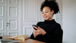 © muse studio - A satisfied female student designer smiles at an Afro-ethnic appearance. Prints a message on the phone in the messenger. In the browser, working on a new work project. Curly black hair.