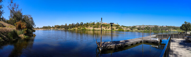 a stunning panoramic shot of vast still blue lake water and mountain ranges filled with lush green plants and trees and luxury homes with an old wooden dock on the lake at Laguna Niguel Regional Park 