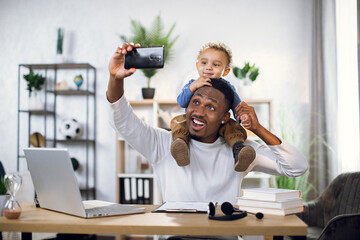 Wall Mural - Handsome young man using smartphone for taking selfie with his cute baby son. Happy father sitting at table with modern laptop and documents.