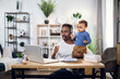 © Sofiia Shunkina - Young african man in headset having video conference on laptop while taking care of his little son at home. Concept of freelance, technology and parenting.