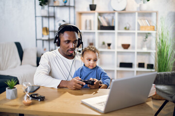 Wall Mural - Afro american father and cute son using modern laptop and wireless joysticks for playing online games. Concept of entertainment, family and leisure time.