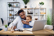 © sofiko14 - African businessman holding his cute son on knees while sitting at table with modern laptop and talking on mobile. Multitasking person at home.