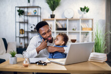 Afro American Man Talking On Mobile, Working On Laptop And Carrying Baby Son. Male Freelancer Sitting At Home Office And Using Modern Gadgets.