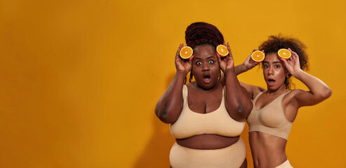 Wall Mural - Two funny young african american women looking surprised at camera, holding halves of ripe juicy orange near their faces, posing isolated over orange background