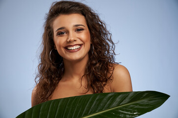 Wall Mural - Portrait of young woman with perfect makeup laughing, hiding chest behind big green leaf. Attractive sincere naked female model with curly hair and big earrings looking at camera isolated on blue.