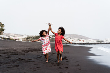 Black twin sisters running on the beach while playing with wood toy airplane - Focus on faces