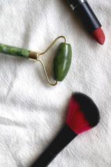 Jade face roller, make-up brush and lipstick on white linen background. Flat lay.