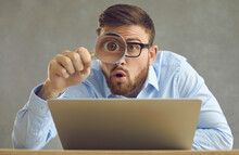 Portrait Of Funny Handsome Nerdy Young Business Man In Glasses Sitting At Office Desk With Laptop Computer, Holding Magnifying Glass And Looking At Something With Big Eye And Surprised Face Expression