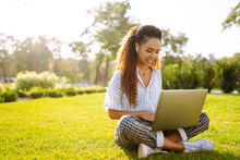 Young Woman Freelancer Sitting On Green Grass With Laptop. Education Online. Happy Woman With Wireless Headphones Calling On Laptop, Talk By Webcam, Video Conference.