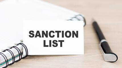 SANCTION LIST text on a business card next to a notebook and a black pen on the table.