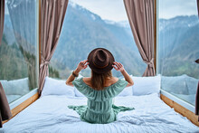 Young Charming Beautiful Boho Chic Brunette Woman Traveler Wearing Bare Shoulder Emerald Dress And Felt Brown Hat Sitting On Bed With Mountain View
