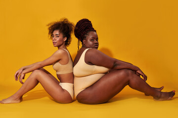 Full length shot of two diverse african american female models in underwear sitting together isolated over yellow background