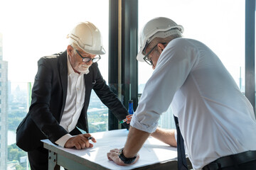 Two engineers discuss their project. They wear white shirt and suit. They also wear white hard hat. They use a pen and paper to share their idea.