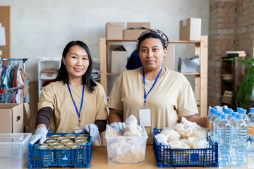 Canvas Print - Two young female volunteers standing by table with free food in plastic boxes