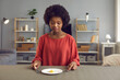 © Studio Romantic - Unhappy African-American woman sits at a table holding a fork and a knife and looks dejectedly at a plate containing only one piece of apple. Concept of hunger, diet and weight loss.
