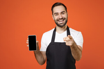 Wall Mural - Young man 20s barista bartender barman employee in black apron white t-shirt work in coffee shop hold mobile cell phone point finger camera on you isolated on orange background Small business startup.