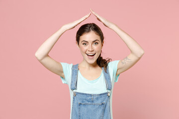 Wall Mural - Young happy caucasian woman in trendy denim clothes blue t-shirt hold folded hands above head like house roof, stay home isolated on pastel pink background studio portrait. People lifestyle concept.