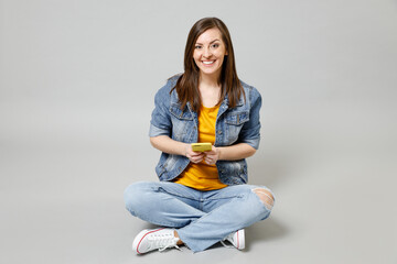 Poster - Full length young happy caucasian woman in casual denim jacket yellow t-shirt use mobile cell phone browsing sitting cross-legged isolated on grey background studio portrait. People lifestyle concept