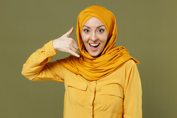 Poster - Friendly young arabian asian muslim woman in abaya hijab yellow clothes do phone gesture like says call me back isolated on olive green background. People uae middle eastern islam religious concept