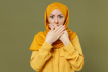 Wall Mural - Shocked scared astonished young arabian asian muslim woman in abaya hijab yellow clothes cover mouth with hands isolated on olive green background People uae middle eastern islam religious concept