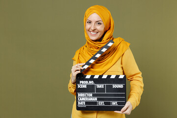 Smiling happy young arabian asian muslim woman 20s in abaya hijab yellow clothes holding classic black film making clapperboard isolated on olive green background. People uae islam religious concept