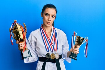 Wall Mural - Beautiful brunette young woman wearing karate fighter uniform and medals holding trophy clueless and confused expression. doubt concept.