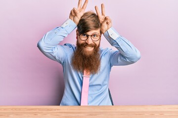 Wall Mural - Young irish redhead man wearing business shirt and tie sitting on the table posing funny and crazy with fingers on head as bunny ears, smiling cheerful