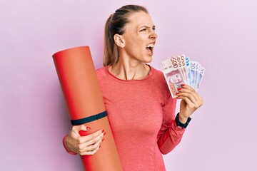 Wall Mural - Beautiful caucasian sports woman holding yoga mat and swedish krona banknotes angry and mad screaming frustrated and furious, shouting with anger. rage and aggressive concept.