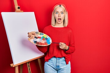 Poster - Beautiful blonde woman standing drawing with palette by painter easel stand afraid and shocked with surprise and amazed expression, fear and excited face.