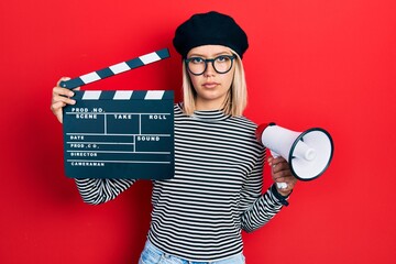 Poster - Beautiful blonde woman holding video film clapboard and megaphone relaxed with serious expression on face. simple and natural looking at the camera.