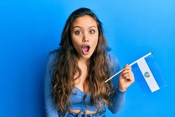 Young hispanic girl holding el salvador flag scared and amazed with open mouth for surprise, disbelief face