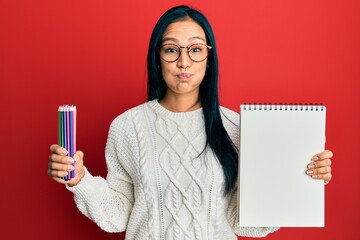 Wall Mural - Beautiful hispanic woman holding canvas book and colored pencils puffing cheeks with funny face. mouth inflated with air, catching air.