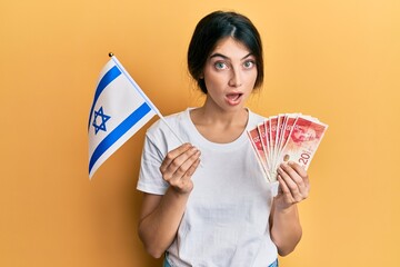 Wall Mural - Young caucasian woman holding israel flag and shekels banknotes afraid and shocked with surprise and amazed expression, fear and excited face.