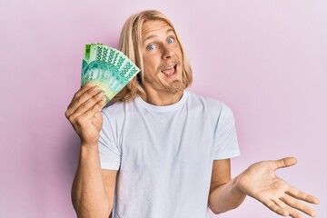 Sticker - Caucasian young man with long hair holding 20000 indonesian rupiah celebrating achievement with happy smile and winner expression with raised hand