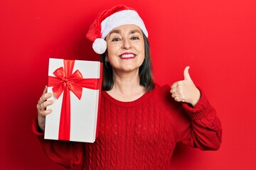 Canvas Print - Middle age hispanic woman wearing christmas hat and holding a gift smiling happy and positive, thumb up doing excellent and approval sign