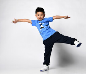 Full body isolated studio portrait of preschool little asian boy wearing blue t-shirt with football player print and sportive pants, sneakers standing on one leg, arms spread wide looking at camera