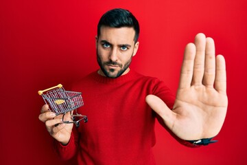 Sticker - Young hispanic man holding small supermarket shopping cart with open hand doing stop sign with serious and confident expression, defense gesture