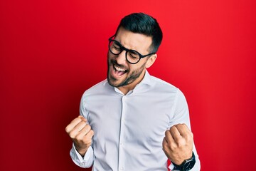 Poster - Young hispanic businessman wearing shirt and glasses very happy and excited doing winner gesture with arms raised, smiling and screaming for success. celebration concept.