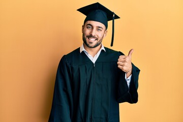 Wall Mural - Young hispanic man wearing graduation cap and ceremony robe smiling with happy face looking and pointing to the side with thumb up.