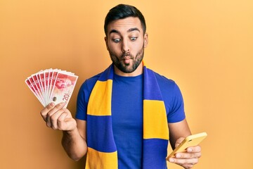 Canvas Print - Young hispanic man football supporter using smartphone holding israel shekels banknotes making fish face with mouth and squinting eyes, crazy and comical.